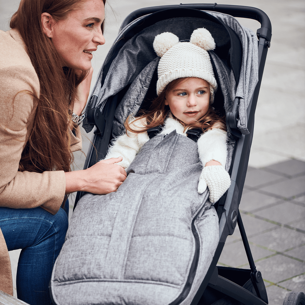 A woman speaks to her child in a stroller with a gray Thule Footmuff.
