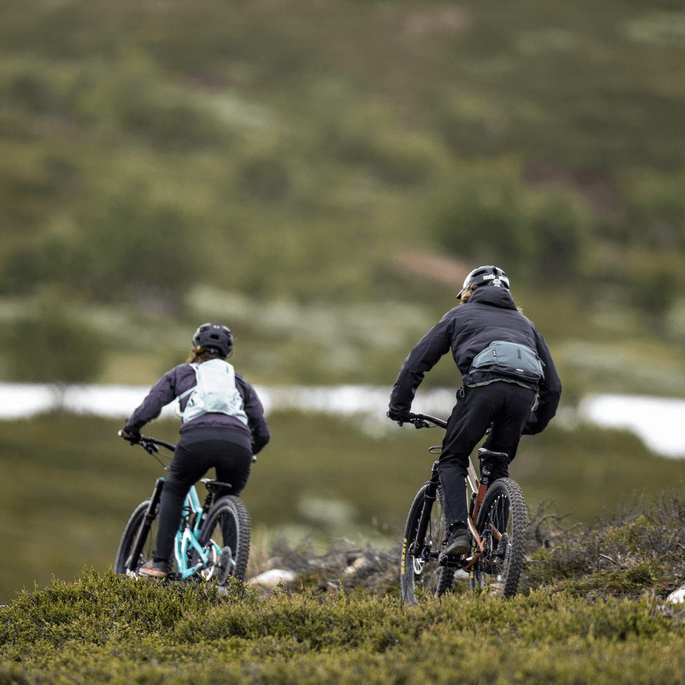Two cyclists bike down a field carrying blue Thule Rail 2L hydration Hip Packs.