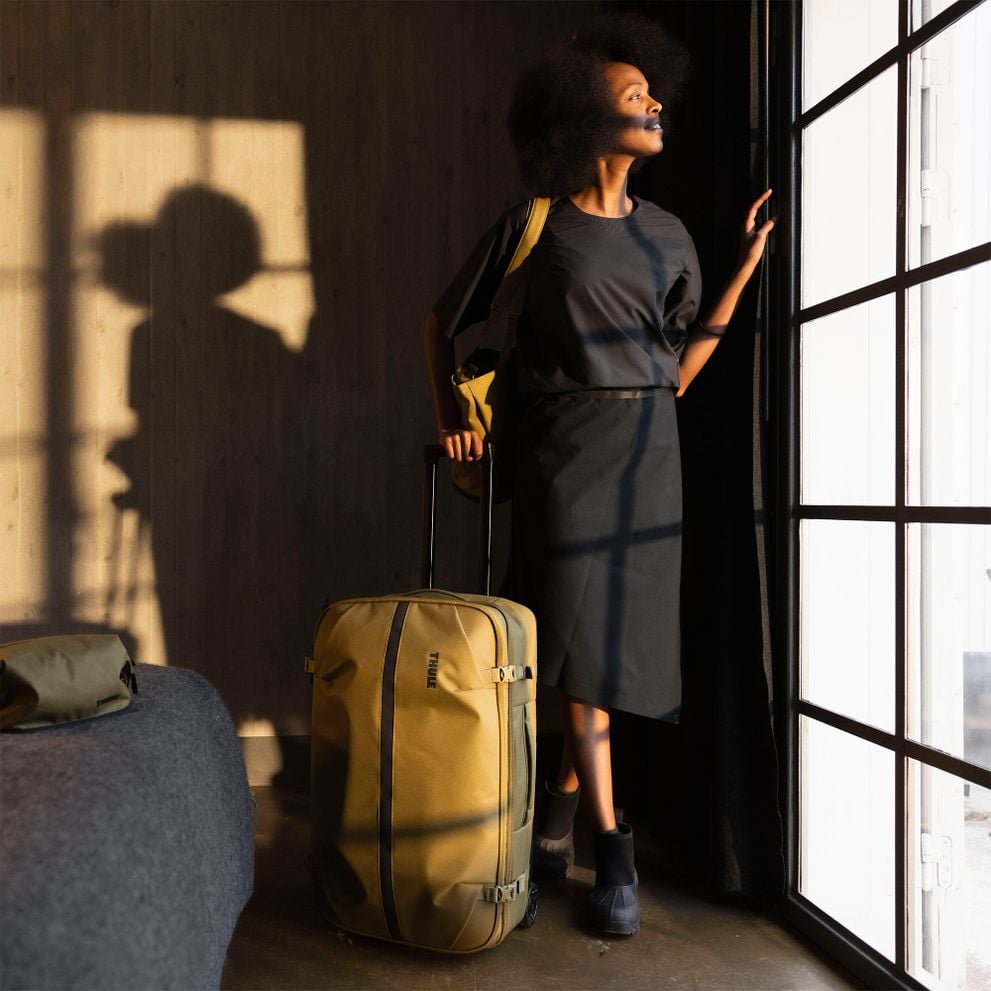 Woman by a window with a Thule Aion rolling suitcase in warm sunlight.