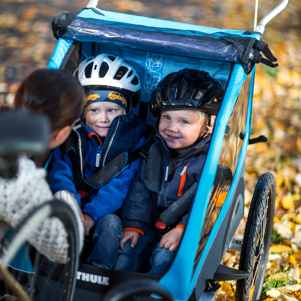 Two kids sit inside a blue A woman bikes down a bike path with a blue Thule Coaster XT kids' bike trailer.