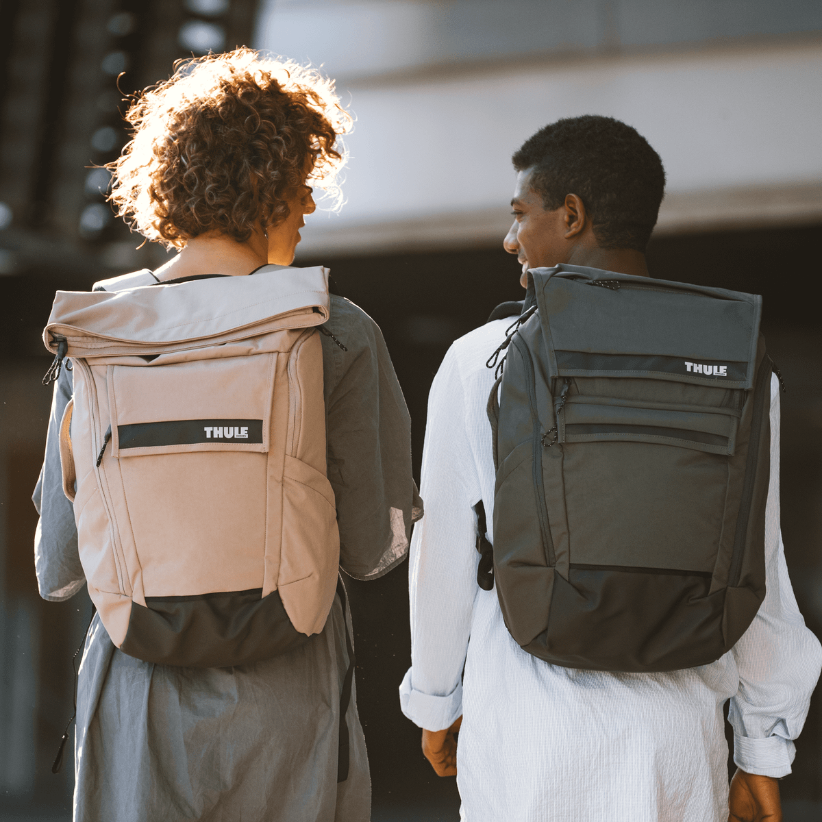 Two friends smile at eachother, carrying green and tan Thule Paramount Backpacks.