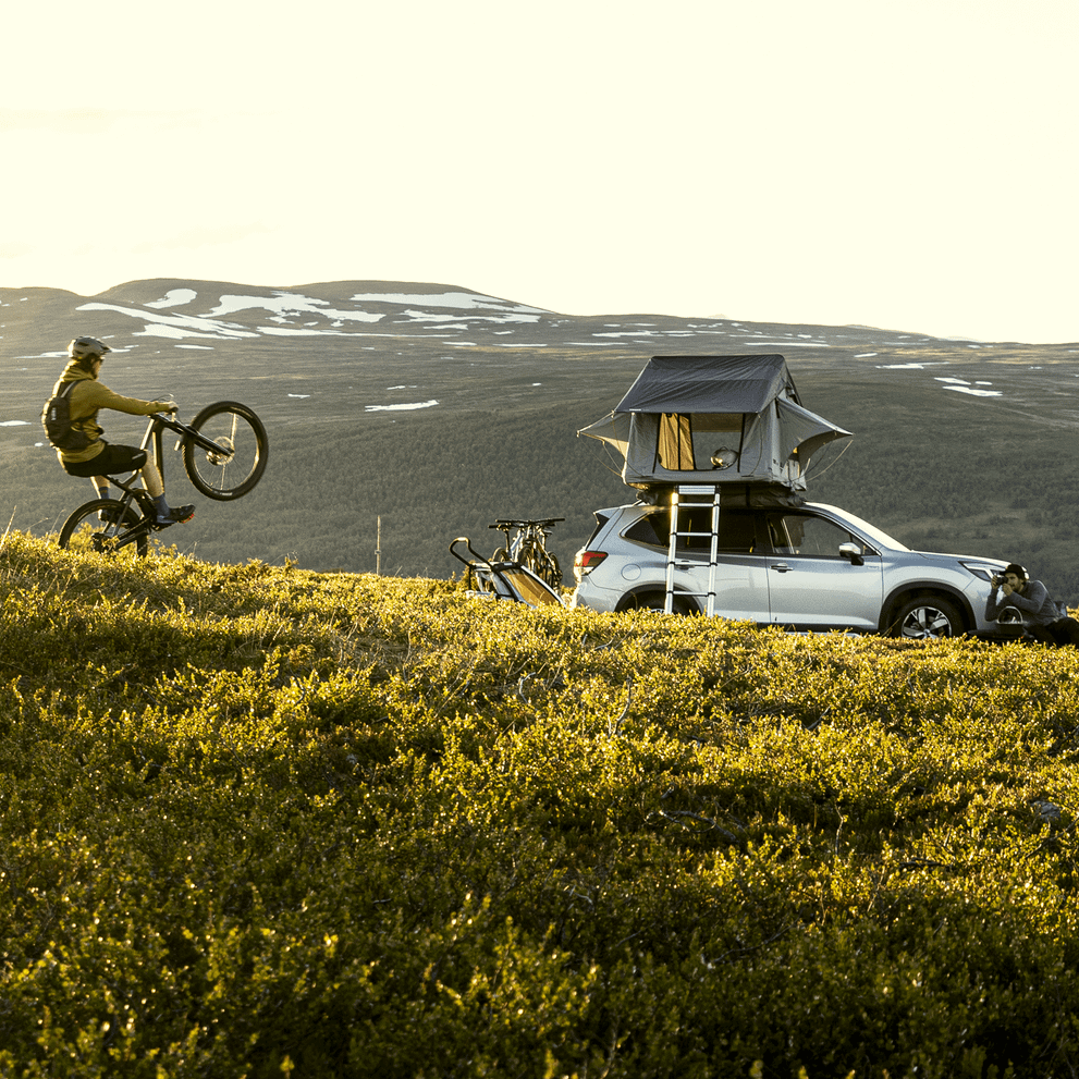 A man is biking towards two cars with outdoor gear. One of the cars has a rooftop tent.