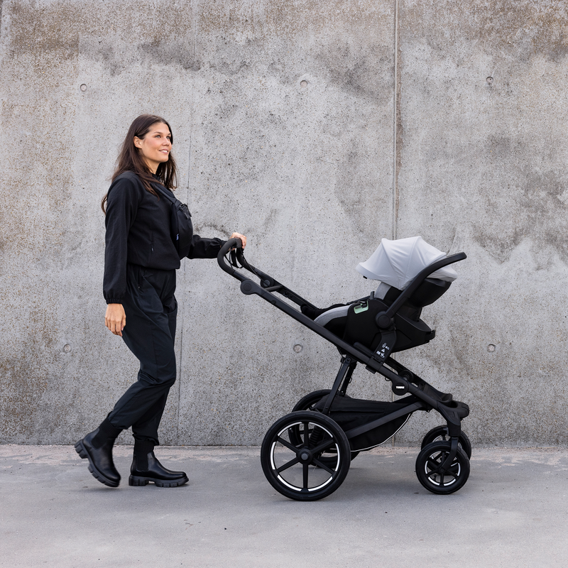 Una donna vestita di nero sta spingendo un passeggino Thule Urban Glide a 4 ruote con un seggiolino auto per neonati Thule Maple in grigio chiaro sopra di esso davanti a un grande muro di cemento.