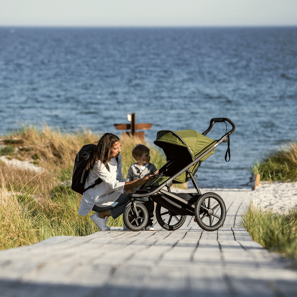 By the beach, a woman leans down to speak to her child in a green Thule Urban Glide 2 all-terrain stroller.