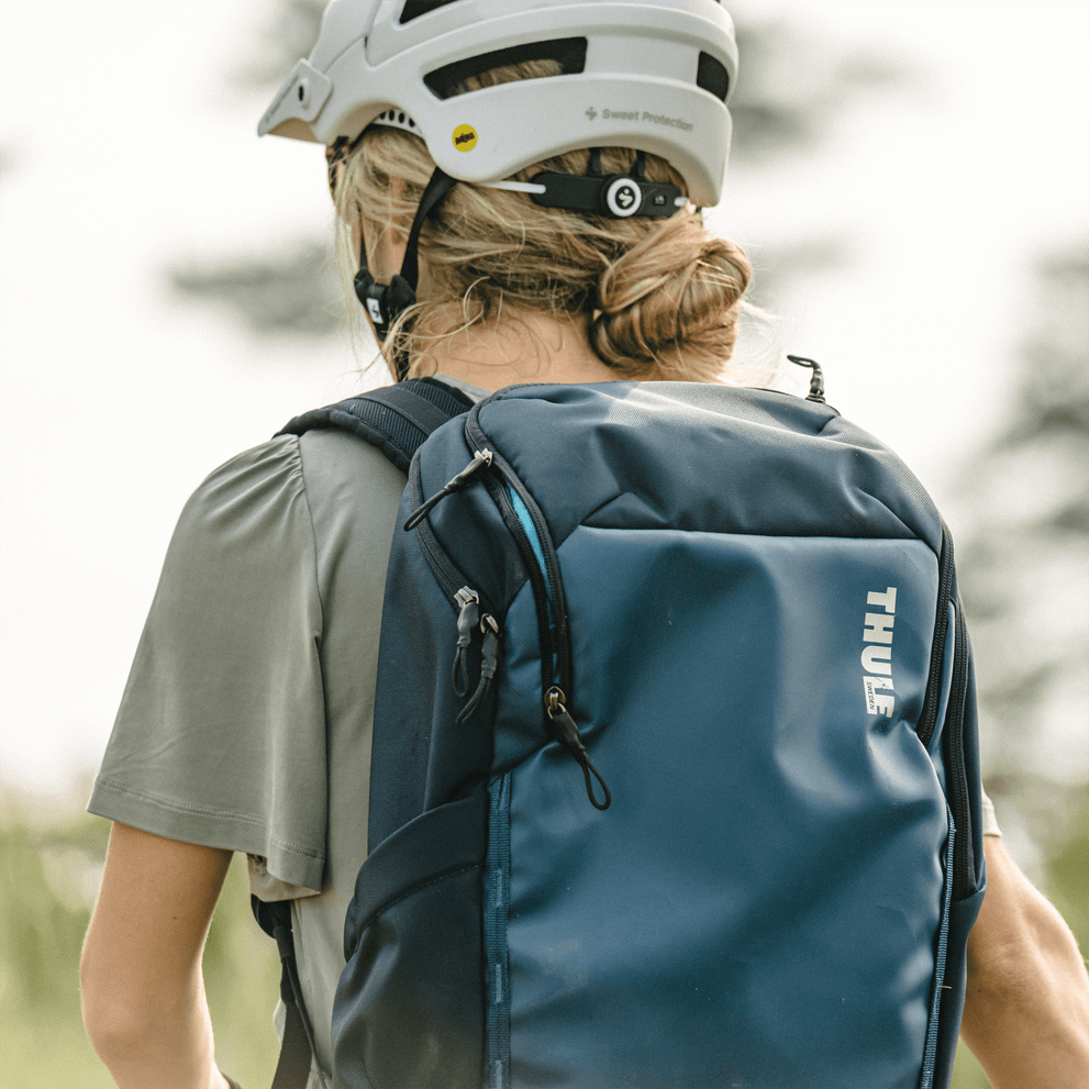 A  woman with a white helmet walks with a blue Thule Chasm backpack.