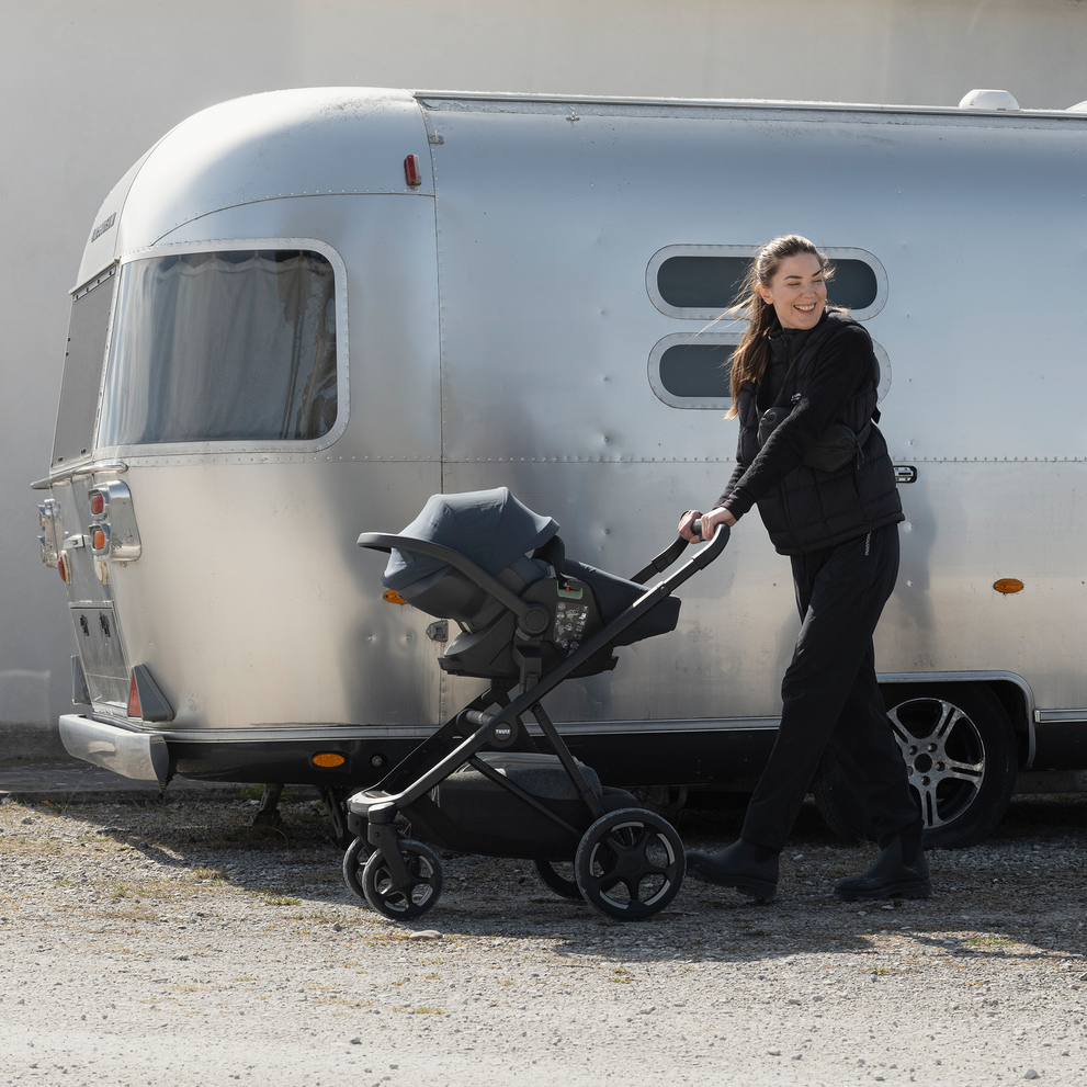Adult pushing a Thule Charm stroller with a Thule Maple infant car seat attached to it beside a camper trailer outdoors.