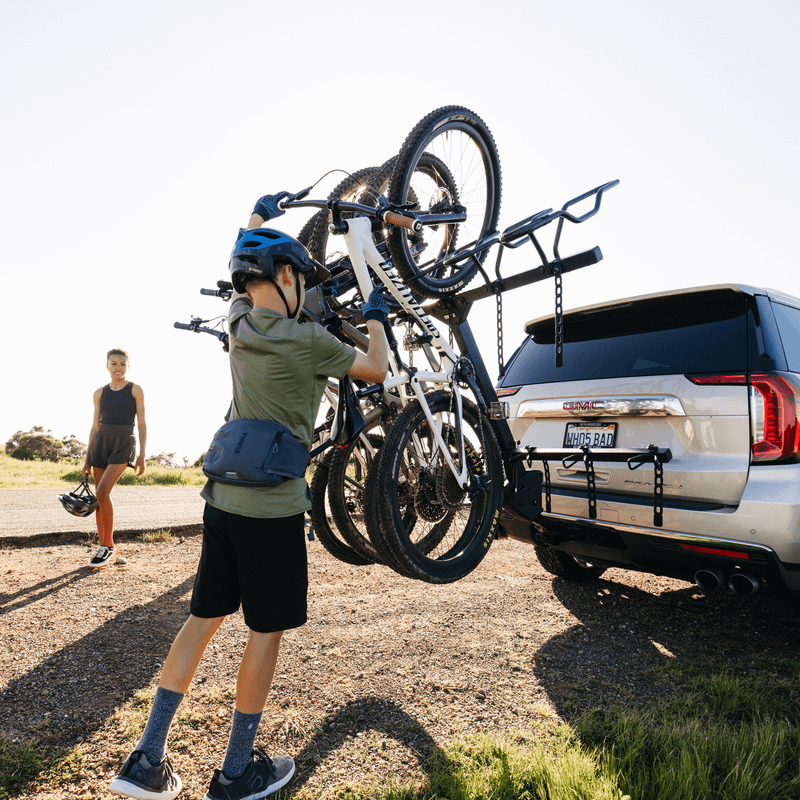 A child is unloading a bicycle from the Thule ReVert hitch bike rack from the back of a silver SUV while one person is watching.