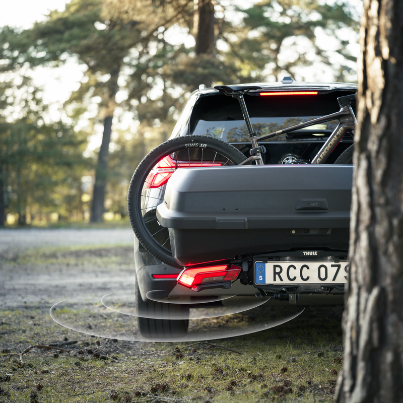 A Thule Epos Parksecure bike rack with active parking sensors and Thule Santu towbar cargo carrier, mounted on the back of a black car carrying one bike and parked in an outdoor setting.