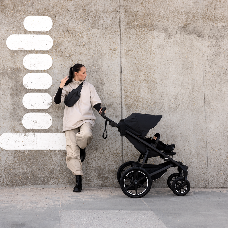A woman leans against a concrete wall with a black Thule Urban Glide 3 four-wheel all-terrain stroller.