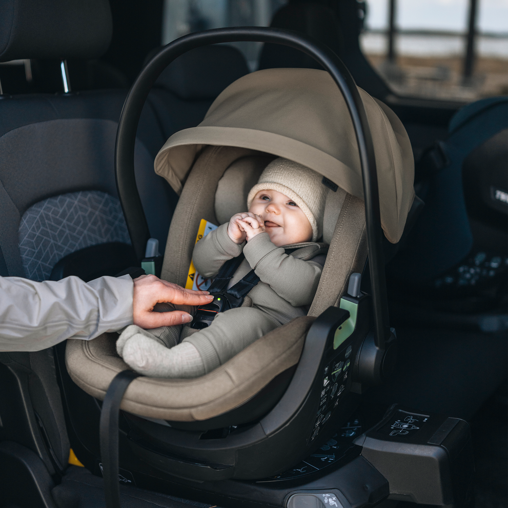 Baby sitting in a rear-facing Thule Maple Connect infant car seat inside a vehicle while an adult adjusts the harness.