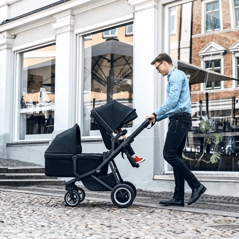In a town street, a father pushes his children in a double stroller with a Thule Sleek Bassinet.
