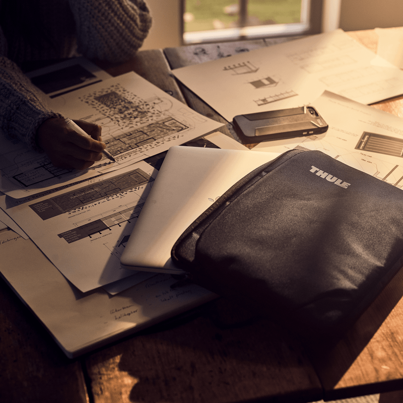 A black Thule Subterra MacBook Sleeve 13" is on a table where a  woman sketches on different papers.