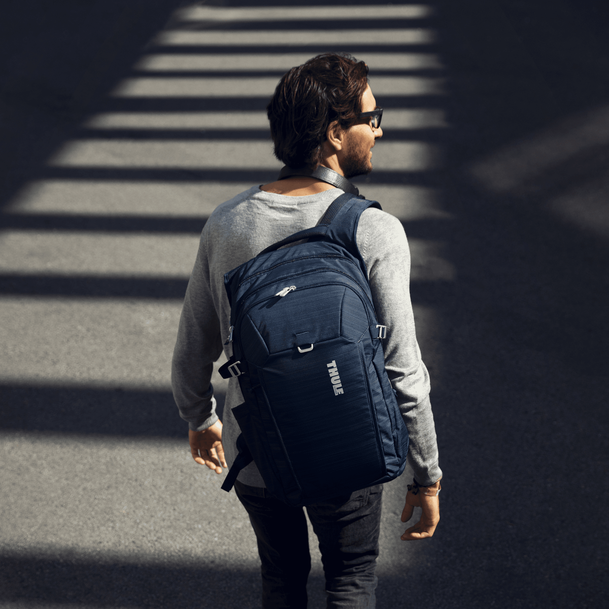 A man crosses a street carrying a blue Thule Construct backpack.