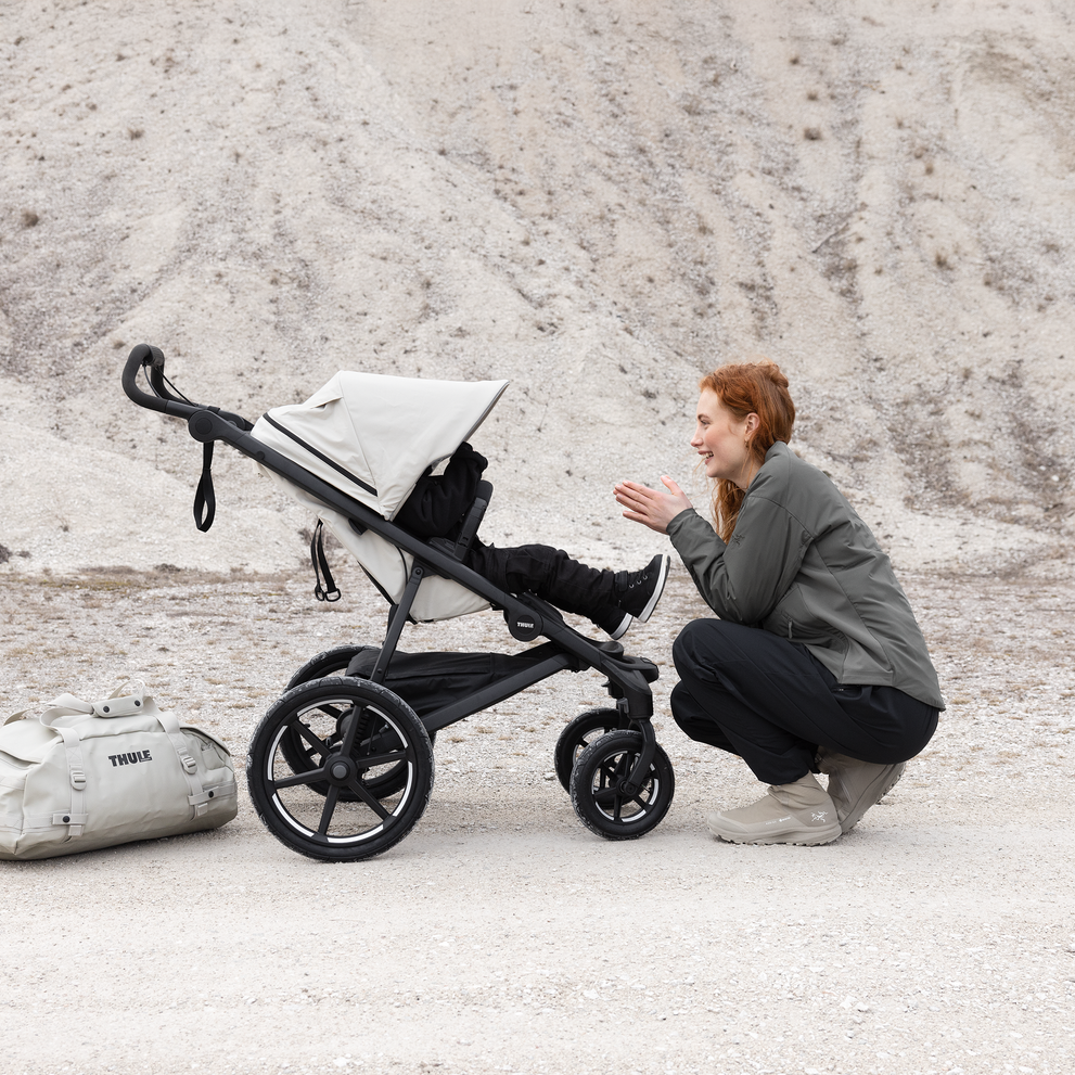 A woman squats in front of a soft beige Thule Urban Glide 4-wheel all-terrain stroller.
