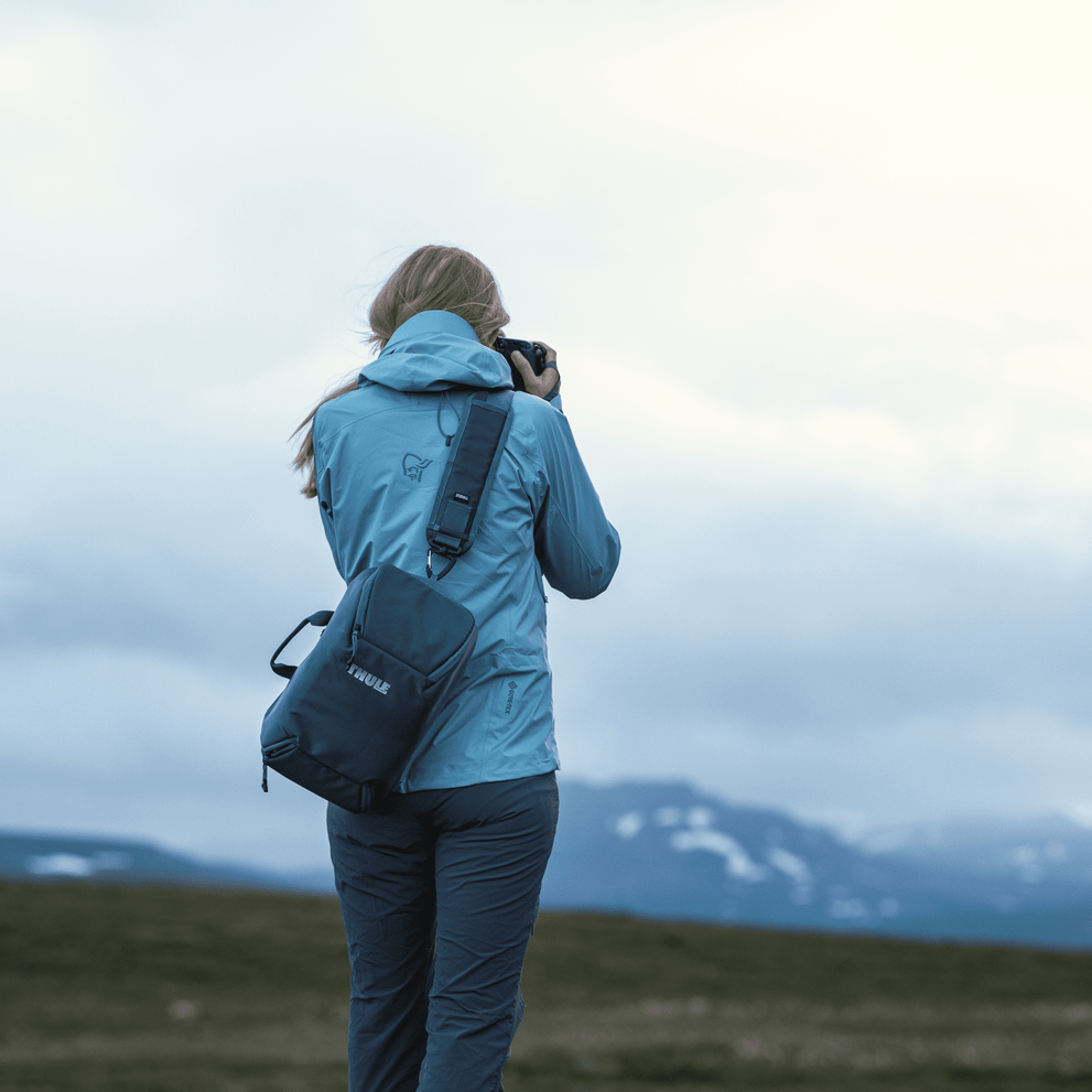 A woman takes a photo of mountains in the distance carrying a blue Thule Covert DSLR camera backpack.