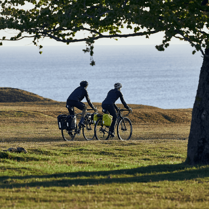 A couple bike by the ocean, with bikes that have black and yellow Thule Shield 25L bike panniers.