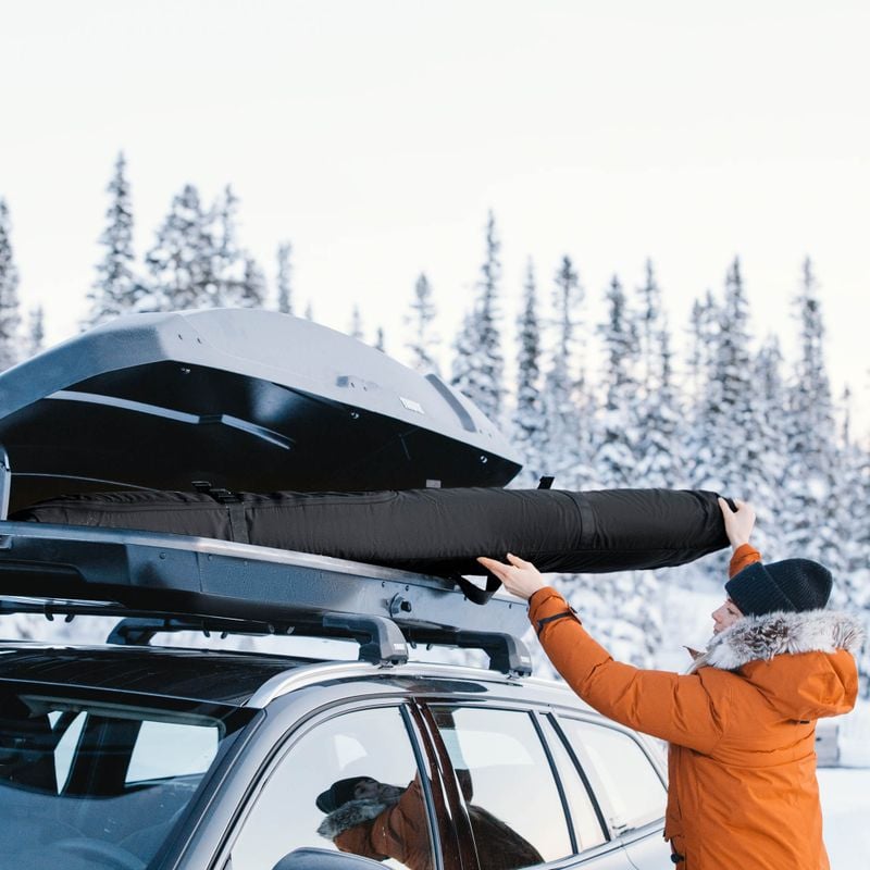 A woman in an orange ski jacket in a snowy setting loads a Thule RoundTrip snowboard bag into the roof box of a car.