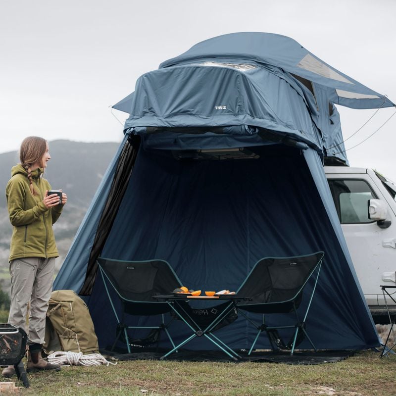 A man and a woman stands in front of a car with a roof top tent that has an annex