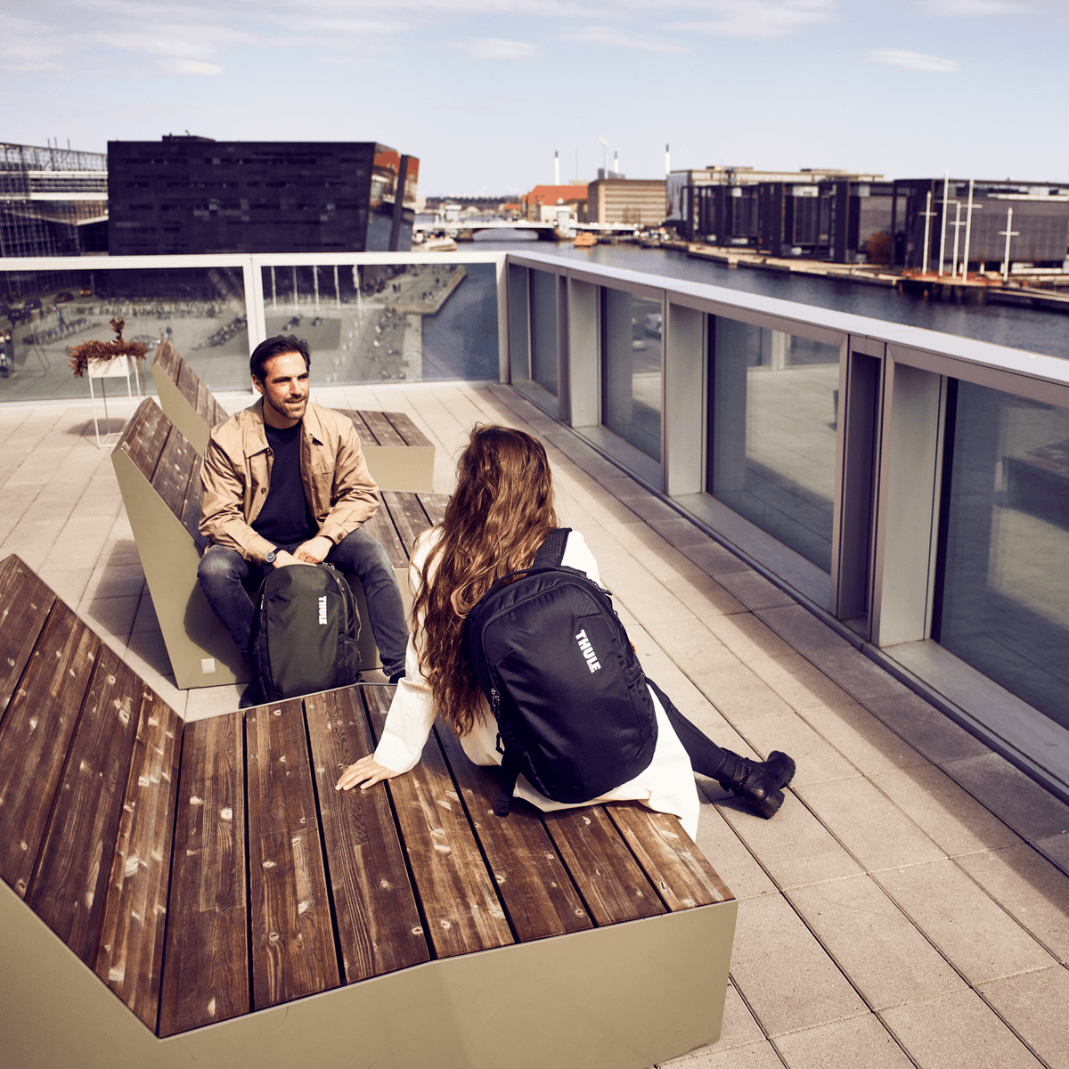 On a terrace overlooking the city, two people sit on a bench talking with Thule Subterra backpacks.