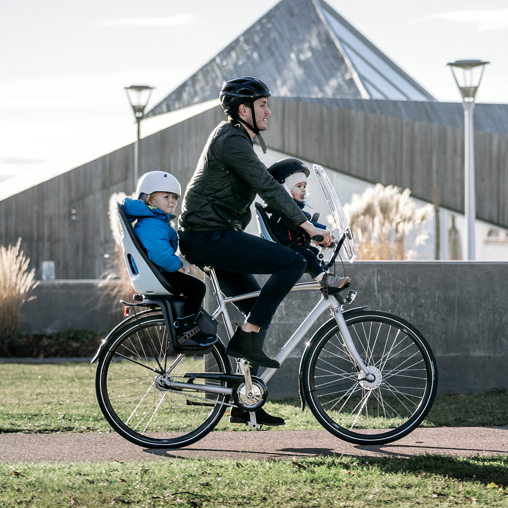 A father bikes down a bike path with two kids in the rear and front Thule Yepp Nexxt Mini child bike seat.