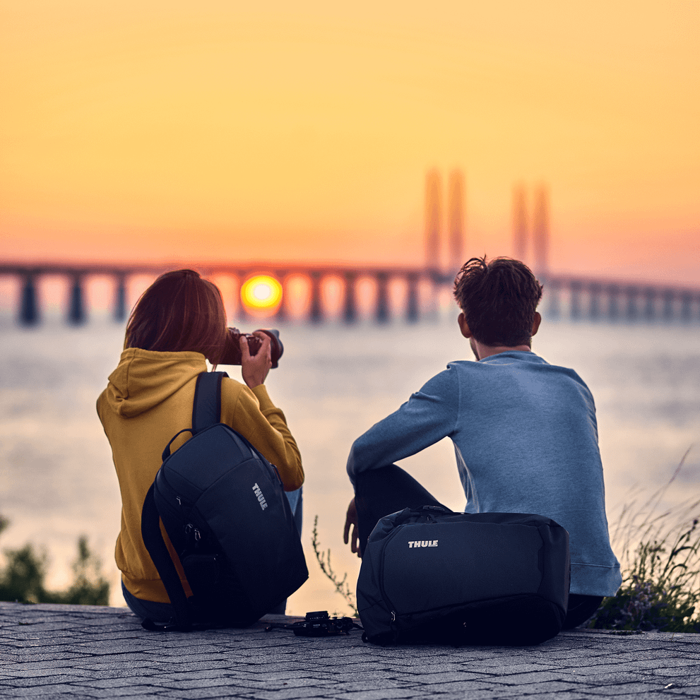 A couple with black Thule EnRoute Medium DSLR camera backpacks take a picture of the sun setting over a bridge.