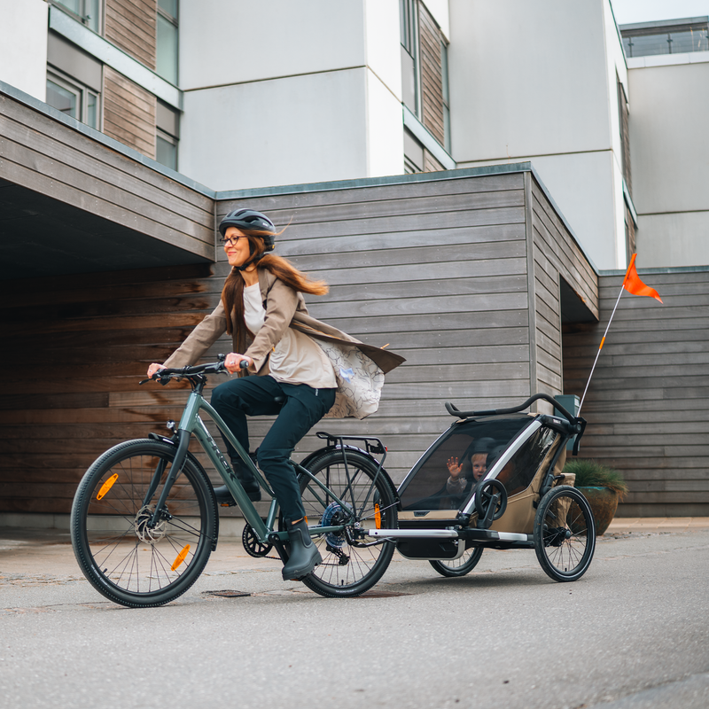 Woman riding a bike while towing two children in a Thule Chariot Cross 2 bike trailer on a city street.