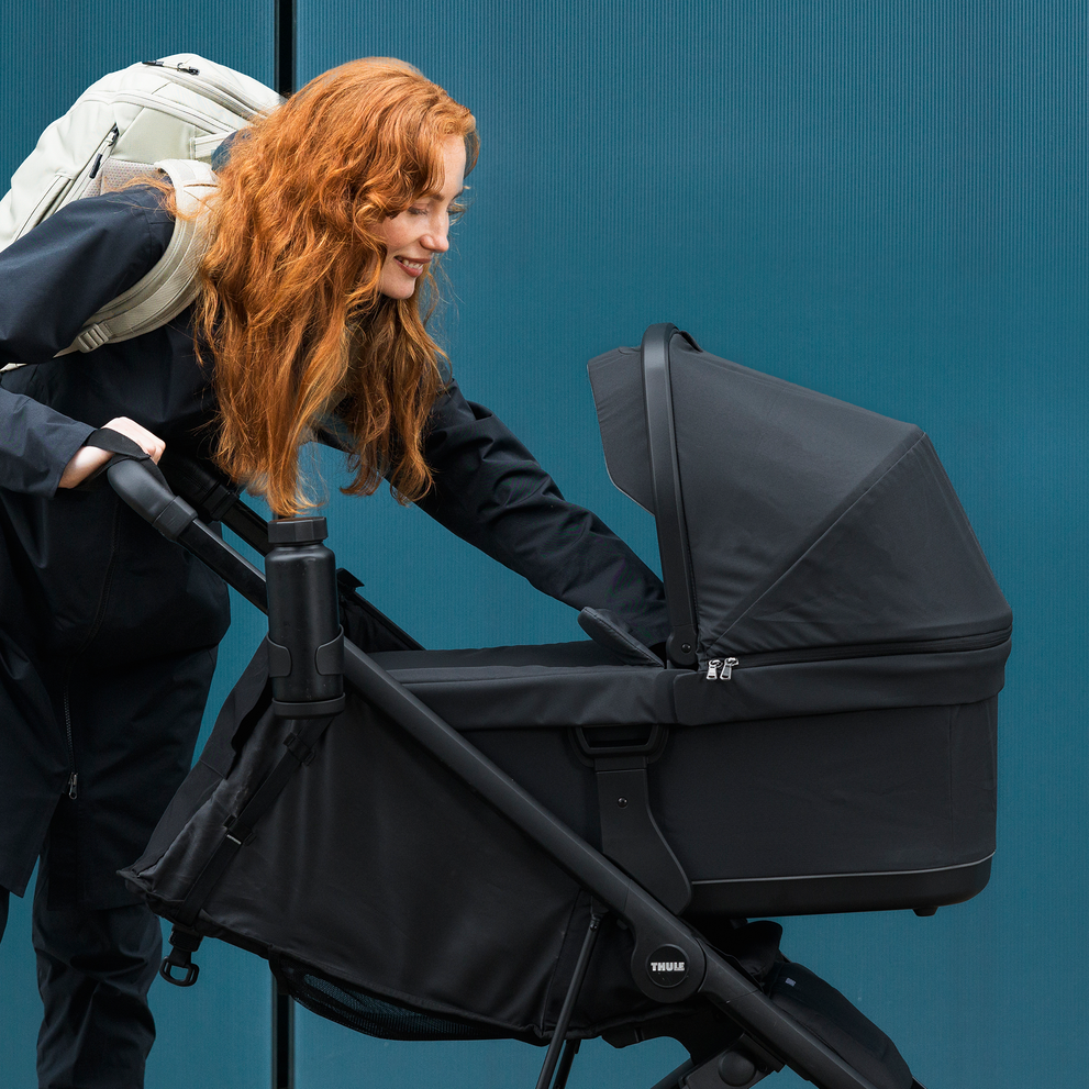 A woman leaning into the Thule Spring 2 stroller with a black bassinet in front of a blue wall.