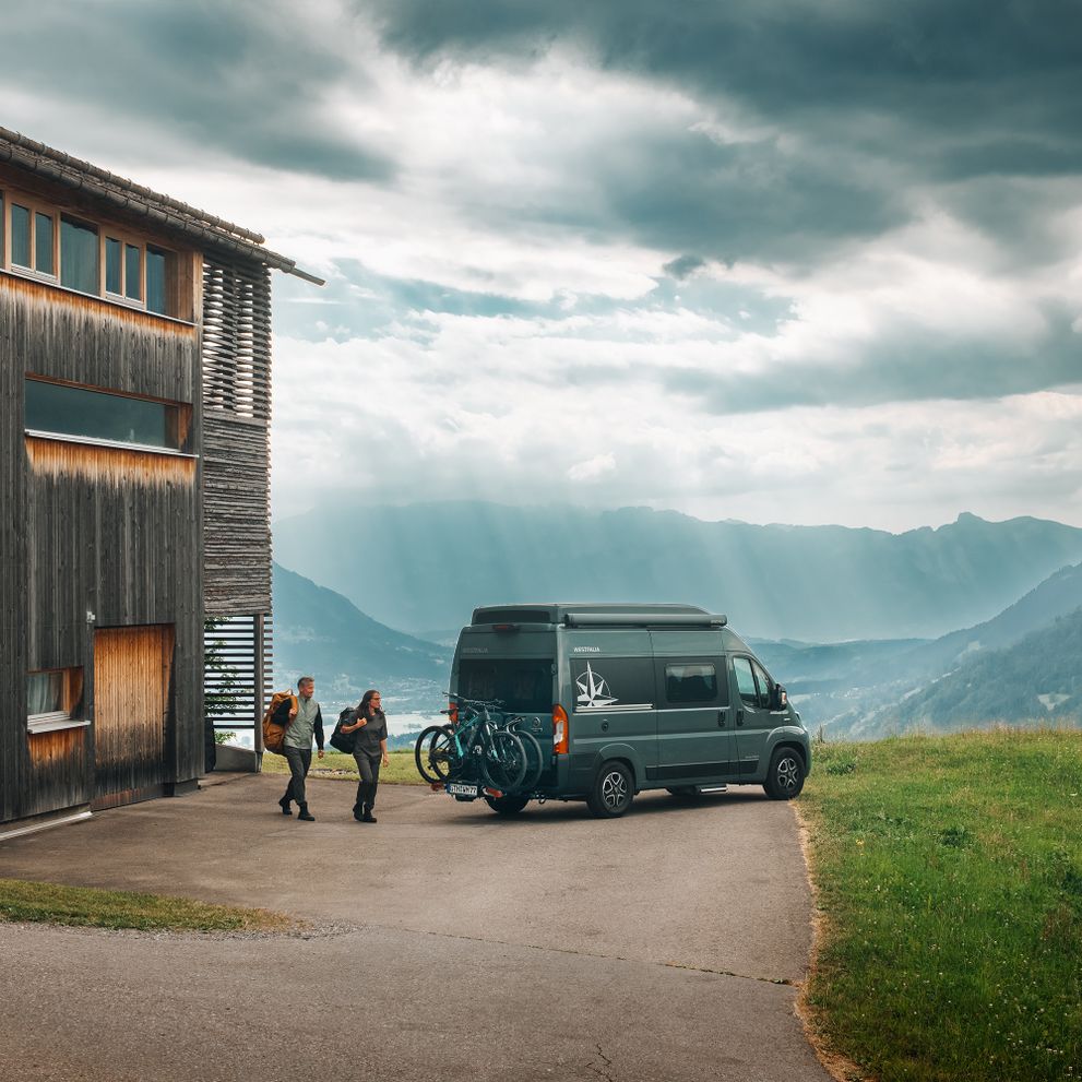 Two people walk towards a gray van with a Thule Veloswing van ebike carrier parked in the mountains.
