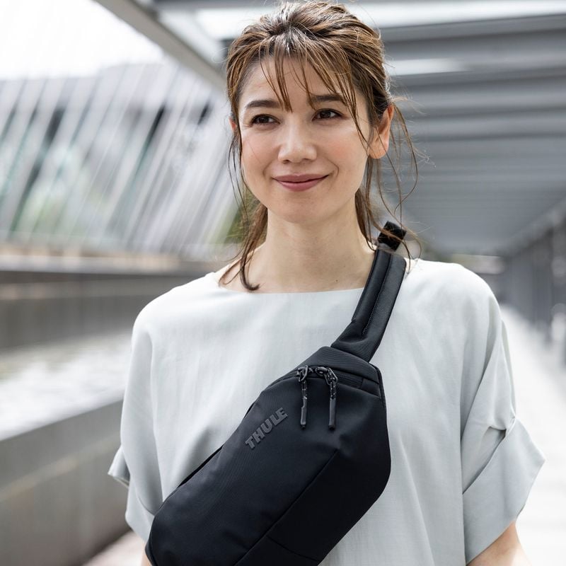 A woman in white looks off in the distance wearing a black Thule Subterra sling bag over her shoulder.