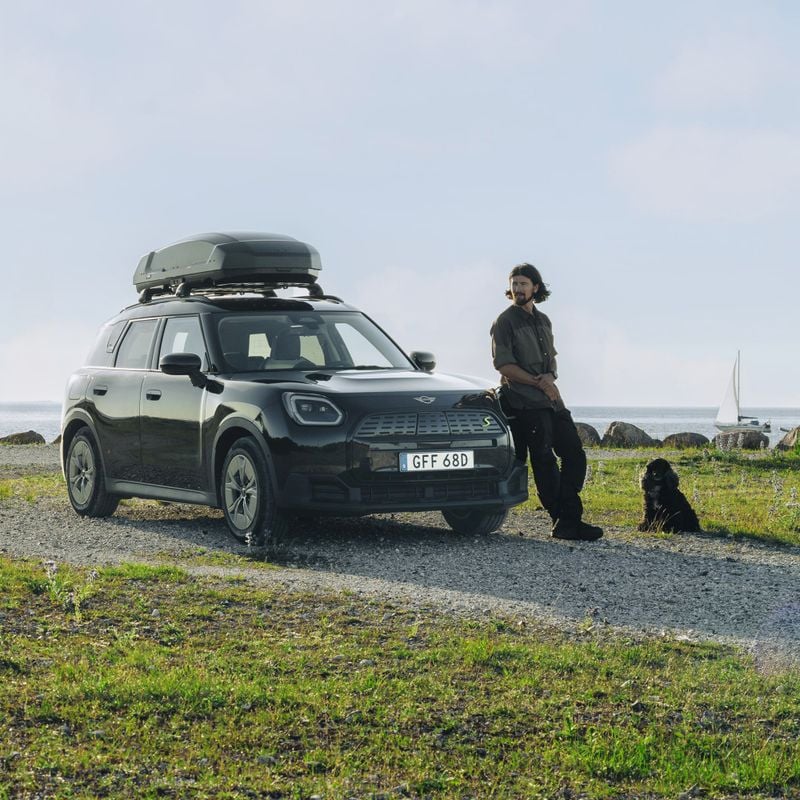 A man and a dog is standing next to a car with a Thule Force 3 rooftop cargo box on its roof, parked on a gravel path by the sea, with clear skies in the background.