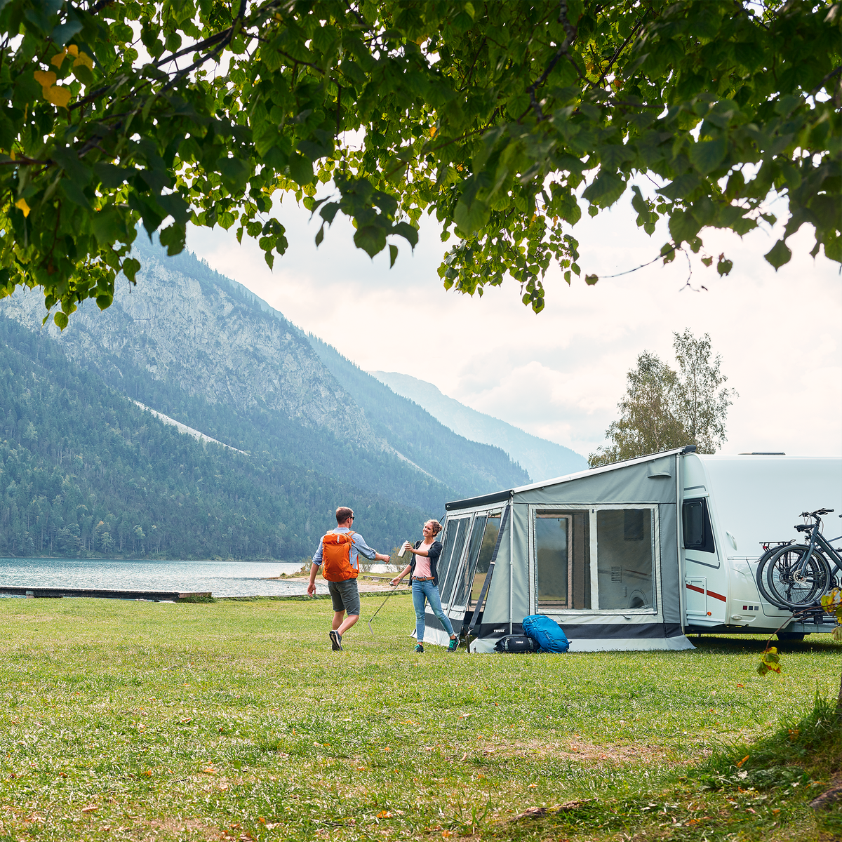 A motorhome parked by a mountain and lake and people stand by a Thule Veduta rv awning tent.