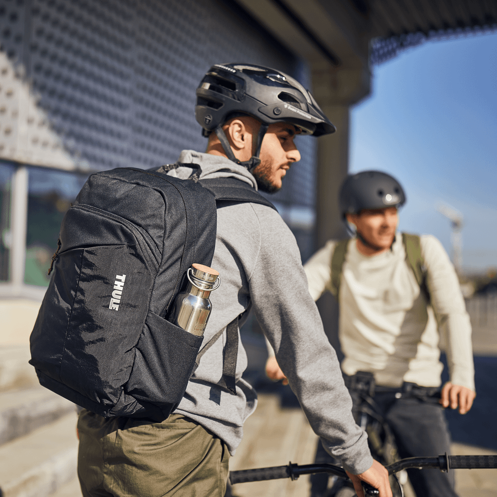 A man holds his bike and a Thule Exeo Backpack talking to another man on a bike.