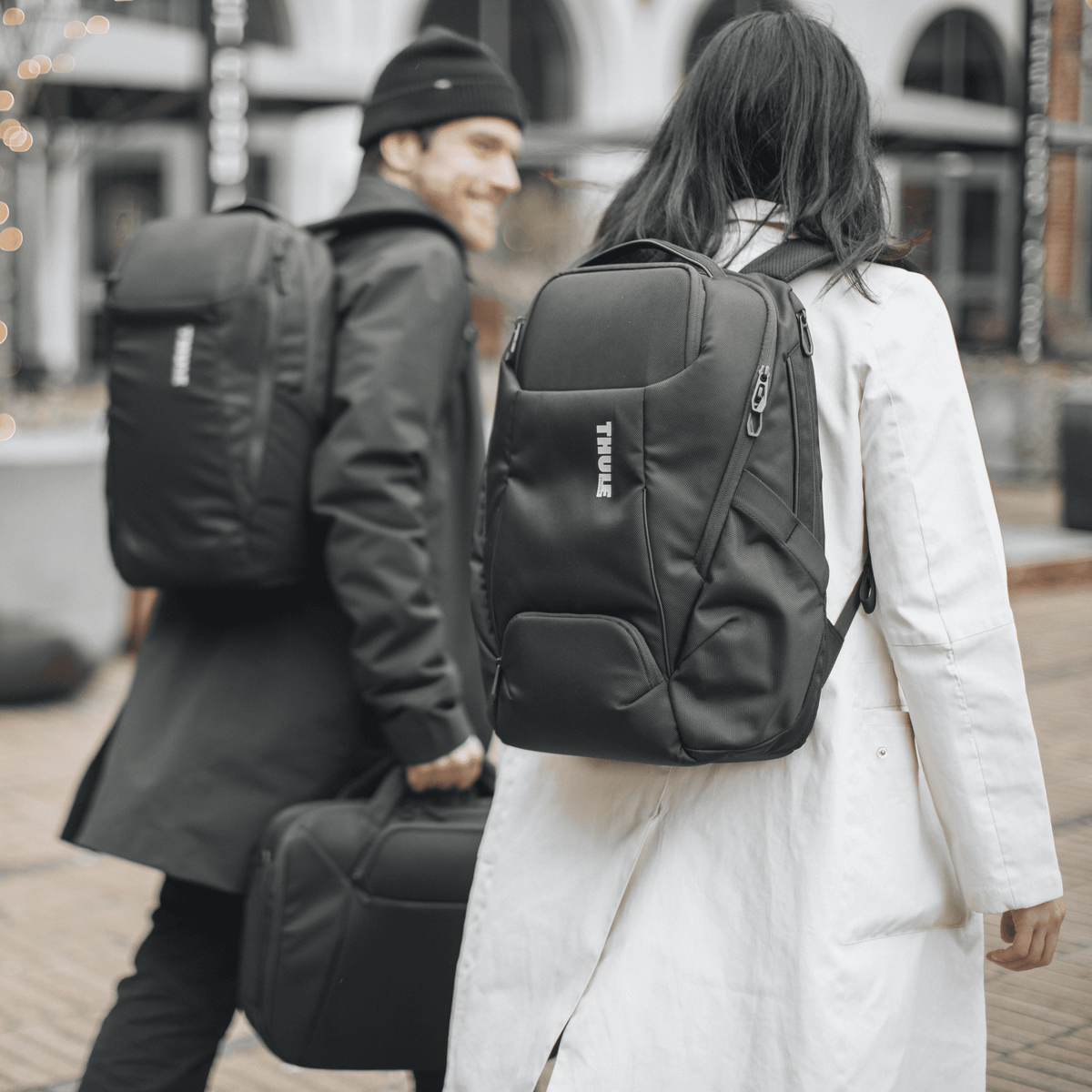A man and a woman walk down a street and chat, carrying black Thule Accent backpacks.