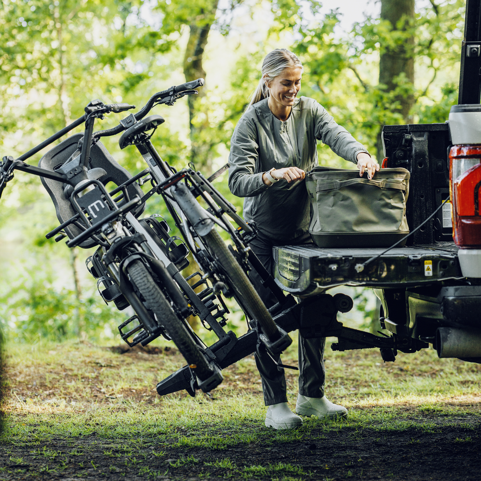 A woman is unloading gear at the back of a truck in an outdoor setting with the Thule Vero hitch bike rack tilted own with two heavy e-bikes.