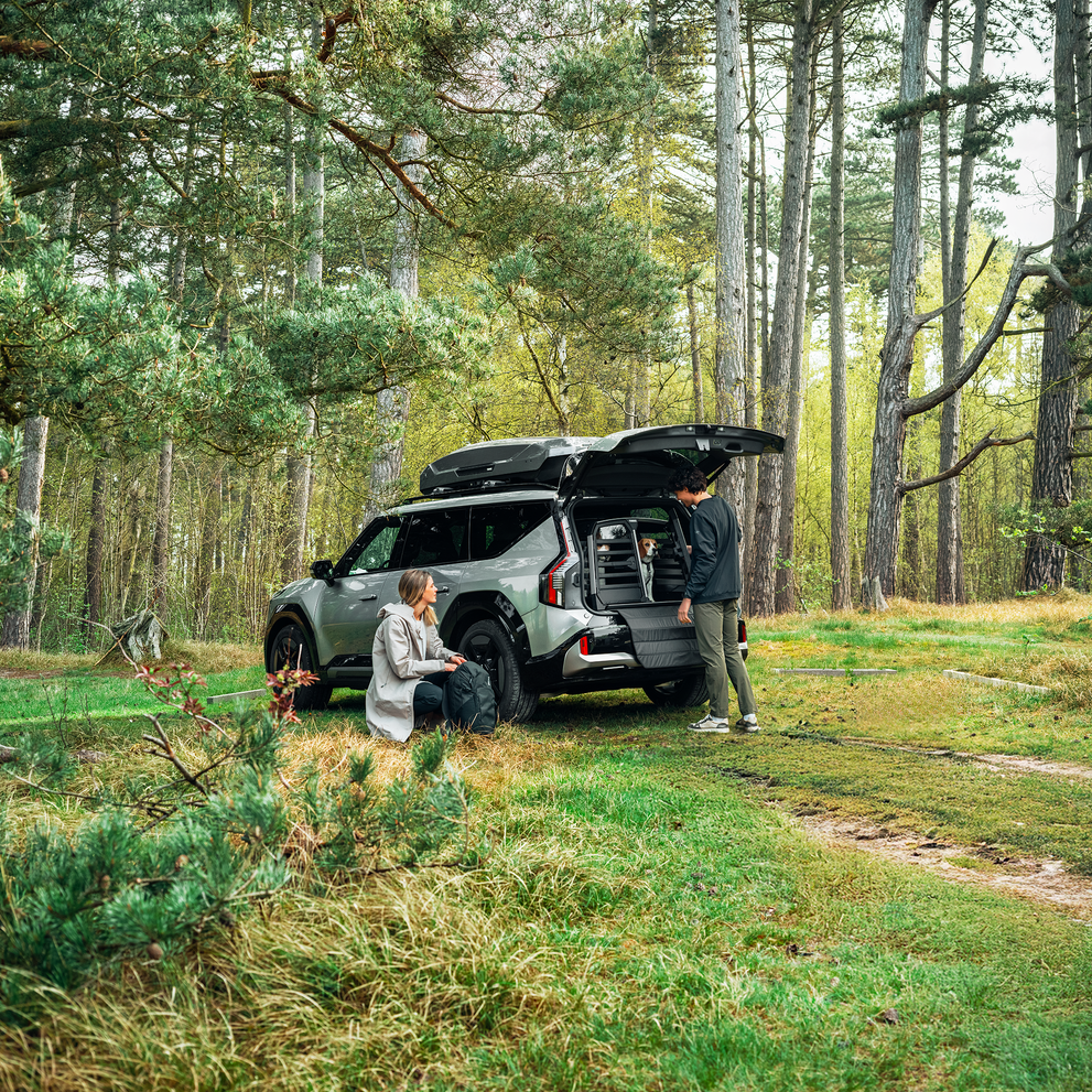 A couple and their dog with their SUV at a forest trailhead, with the Thule Allax double dog car crate visible in the trunk.