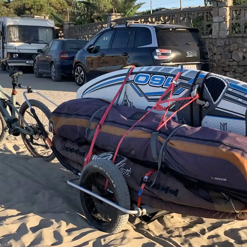 A Reacha Sport Beach bike trailer fully loaded with surfboards and gear, parked near a beach with multiple vehicles in the background.