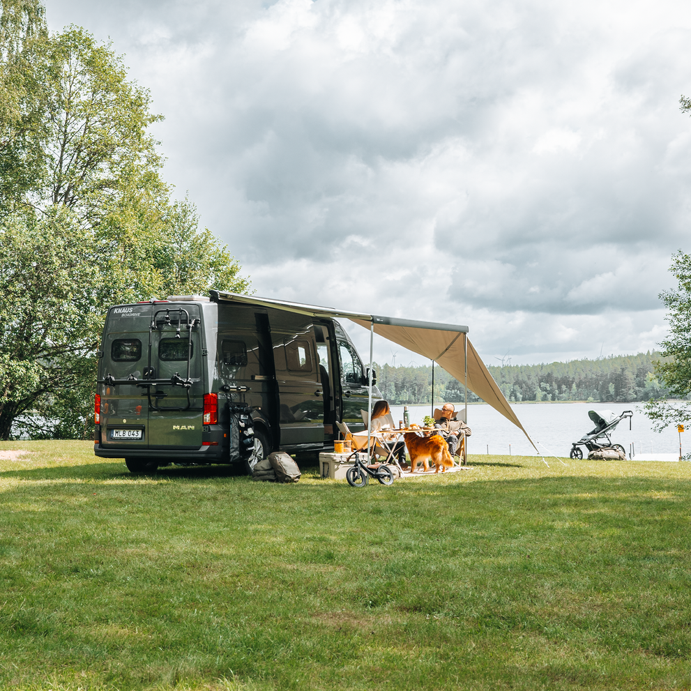 Camper van with extended Thule awning and panels setup by a lake, with people and a dog relaxing under it.