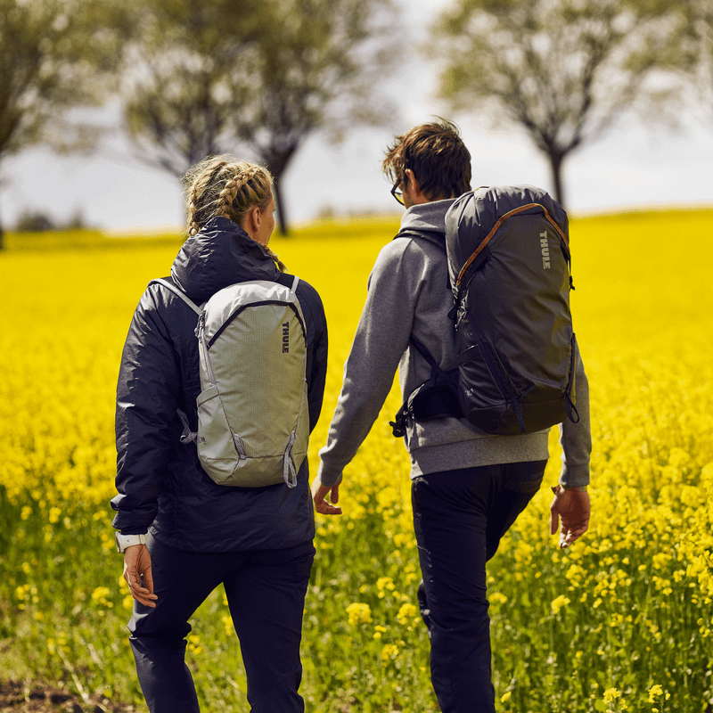 With yellow flowers in the background, a couple walks down a country road with Thule Stir 18L backpacks.