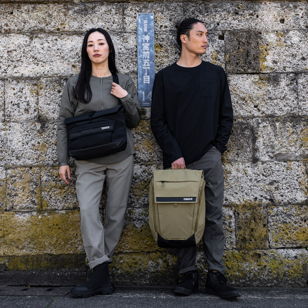 A man and a woman stand against a mossy brick wall with the Thule Paramount backpack 27L.