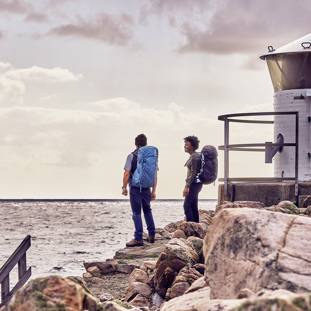 Two people stand by a lighthouse looking out at the ocean carrying Thule Versant 70L backpacks.
