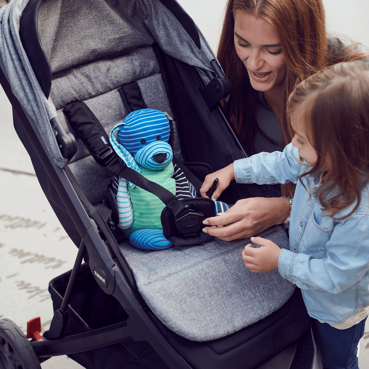 A woman kneels next to her toddler who straps a toy into a gray stroller with a gray Thule Seat Liner.