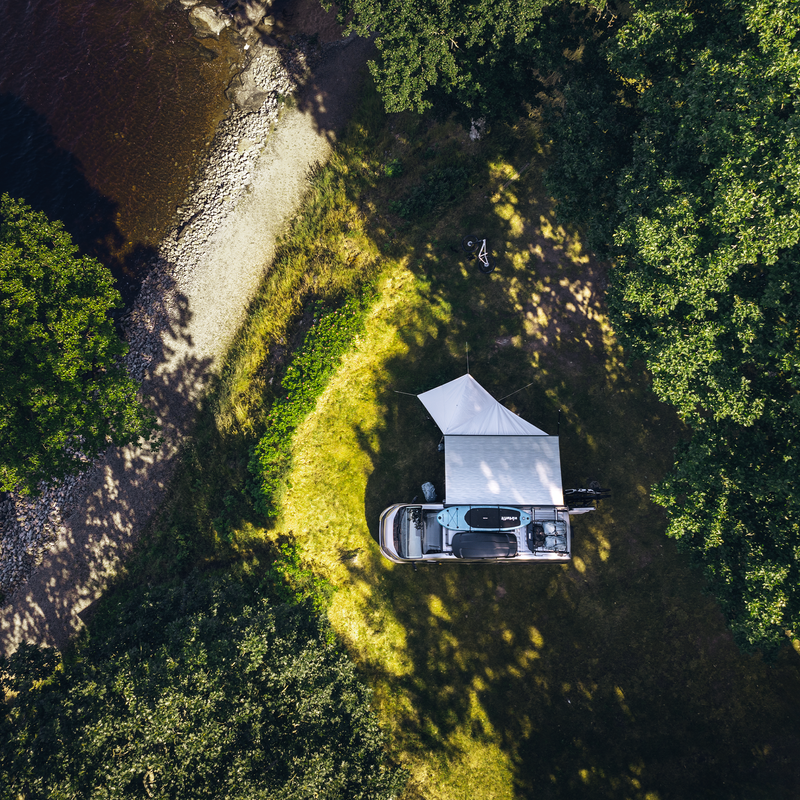 An aerial view of a vehicle parked near a lakeshore, with a Thule Subsola awning extended to create a shaded outdoor space. The setup is surrounded by lush greenery, highlighting the perfect camping spot for adventure seekers.
