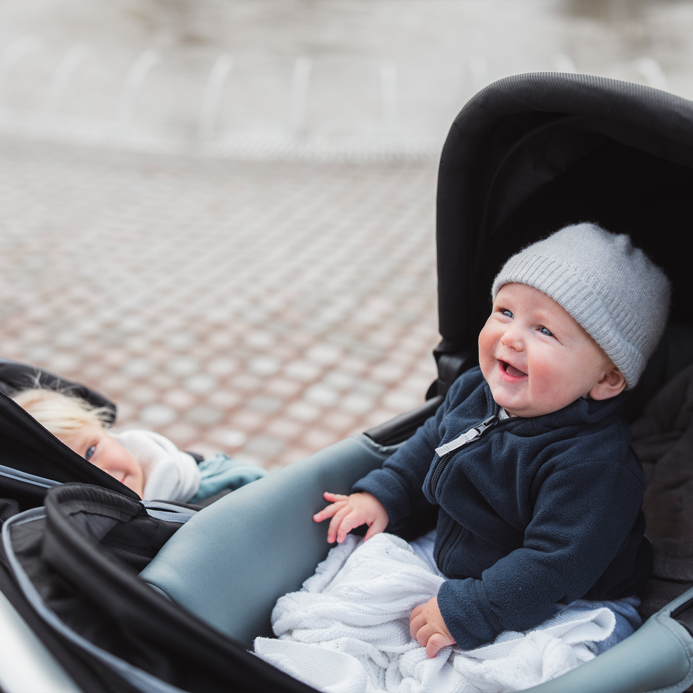 A close-up of two children in a double stroller, one of the kids is in a Thule Urban Glide 2 bassinet.