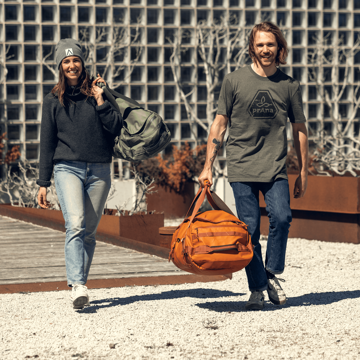 Two people walk in the sunshine with green and orange Thule Chasm duffel bags.