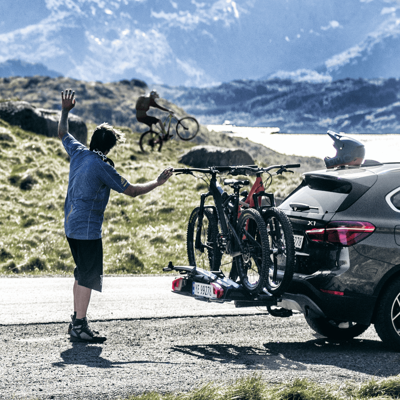 A car with a Thule VeloSpace XT bike rack and bikes is parked next to a gravel road and a man is waving to a friend riding a mountain bike in the distance.