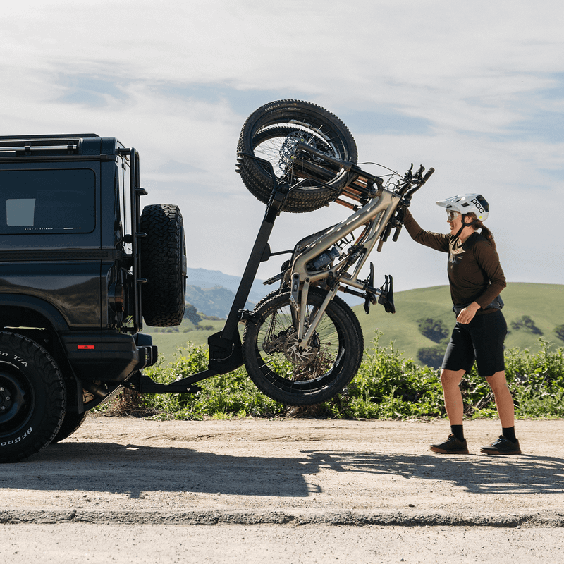 A person is standing behind a black SUV tilting the Thule ReVert hitch bike rack with bicycles mounted on it.  The SUV is parked by the side of the road in a nature setting.
