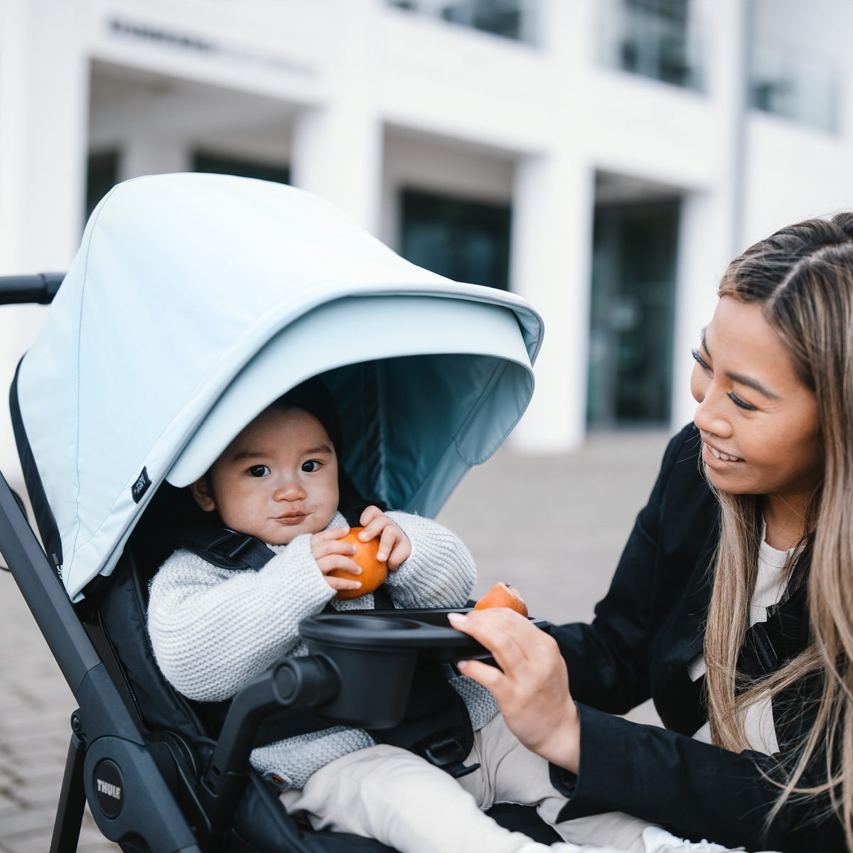 A woman smiles at her baby in a stroller holding an orange placed on the Thule Stroller Snack Tray.