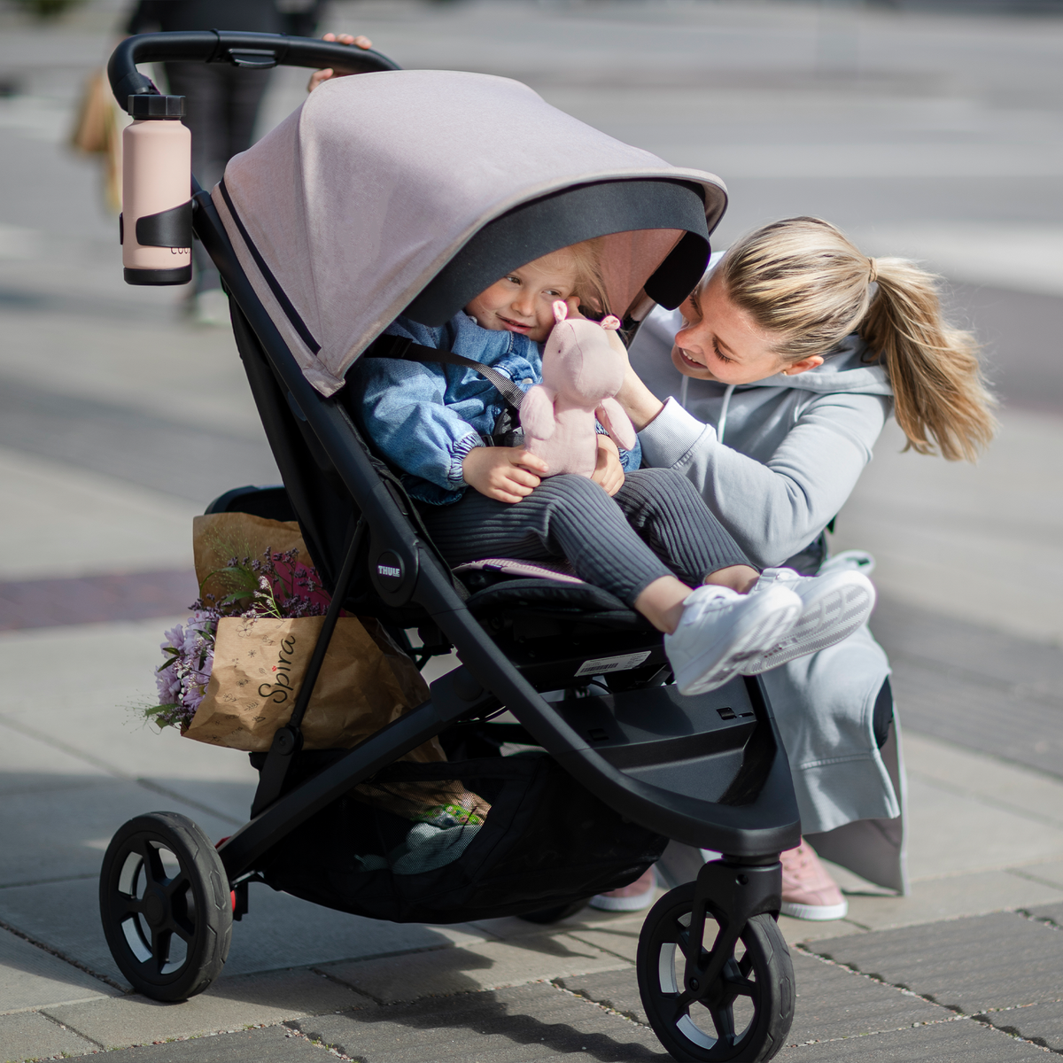 A woman kneels and hands something to her baby sitting in a pink stroller with a pink Thule Summer Seat  Liner.
