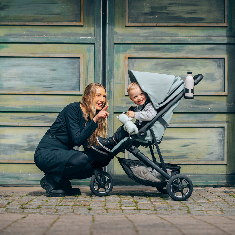 Woman crouching next to toddler in Thule Spring 2 stroller on cobblestone street.