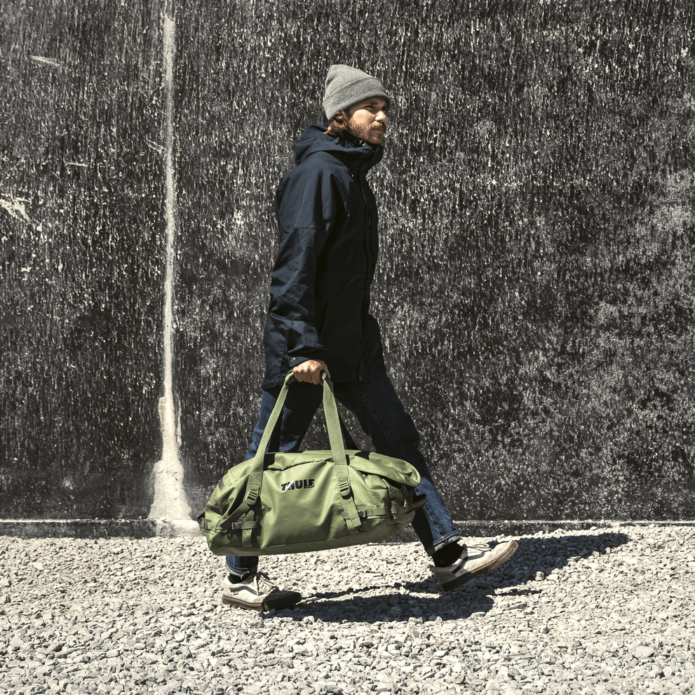 A man walks down a street with a concrete wall holding a green Thule Chasm duffel.